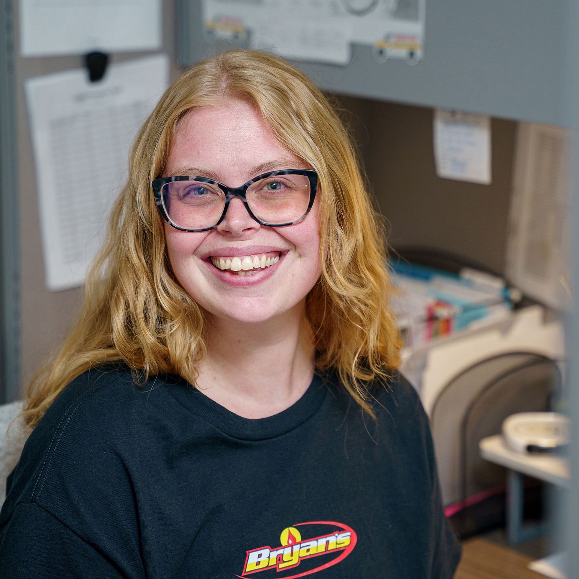 A person with glasses and long hair is smiling, seated in an office setting, wearing a shirt with a colorful logo.