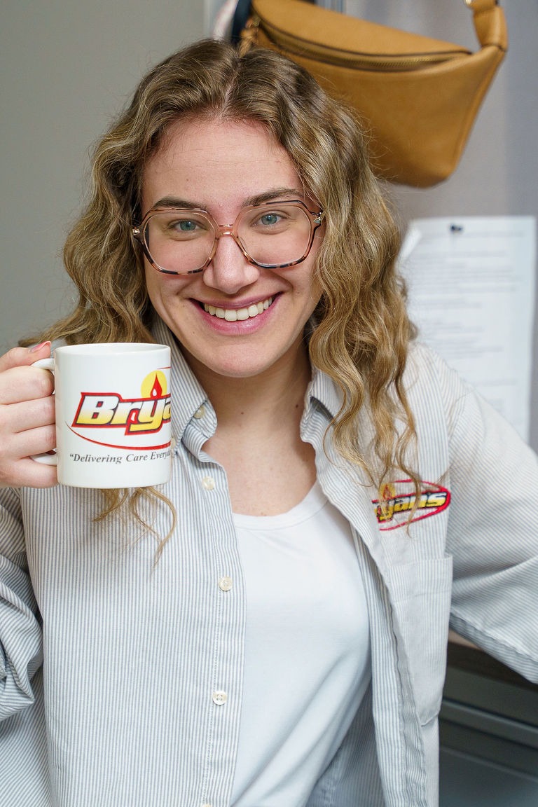 A person with glasses smiles while holding a branded mug with a logo. A handbag is visible in the background.