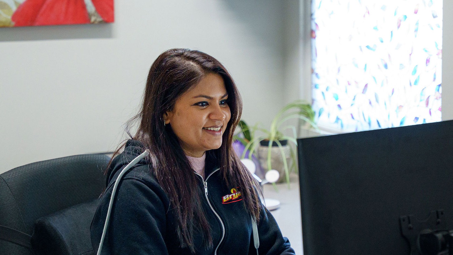 A person is smiling while working at a computer in a brightly lit office with a plant and colorful window.