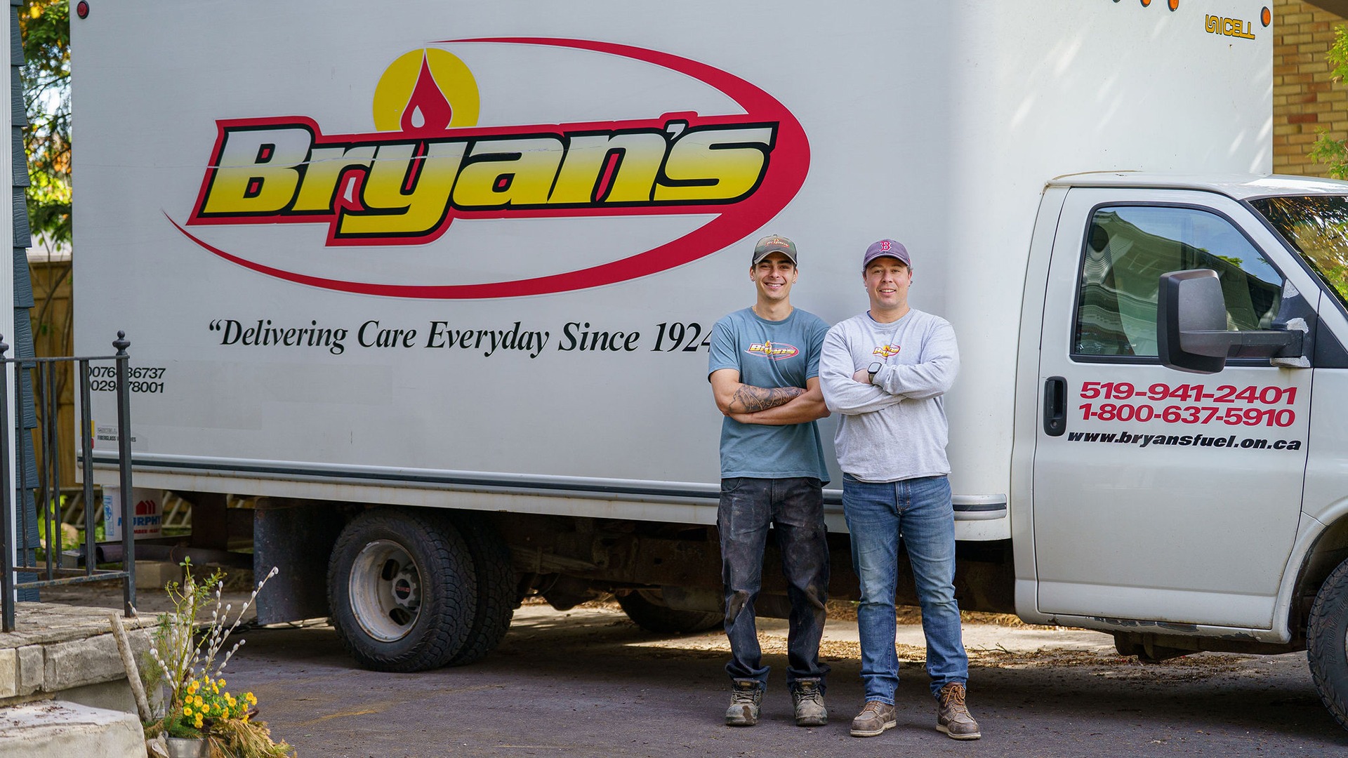Two people stand beside a Bryans Fuel truck, displaying the company logo and contact details. Lush foliage is visible in the background.