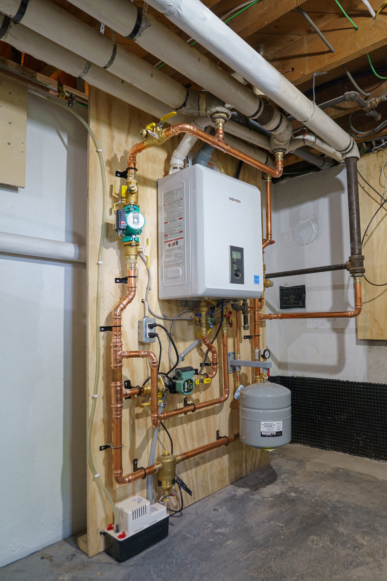 A basement contains a complex network of copper pipes, a tankless water heater, and various plumbing fixtures mounted on a wooden board.
