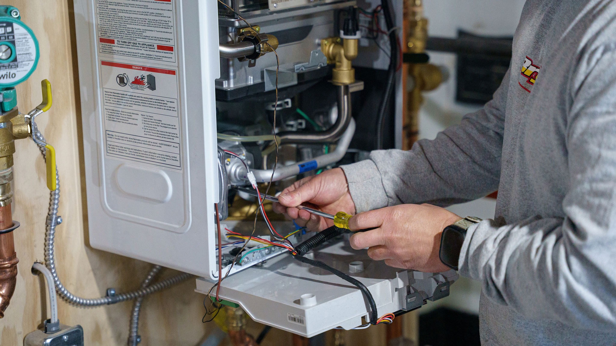 A person is repairing a boiler, using a screwdriver to adjust internal components. Metal pipes and electrical wires are visible.
