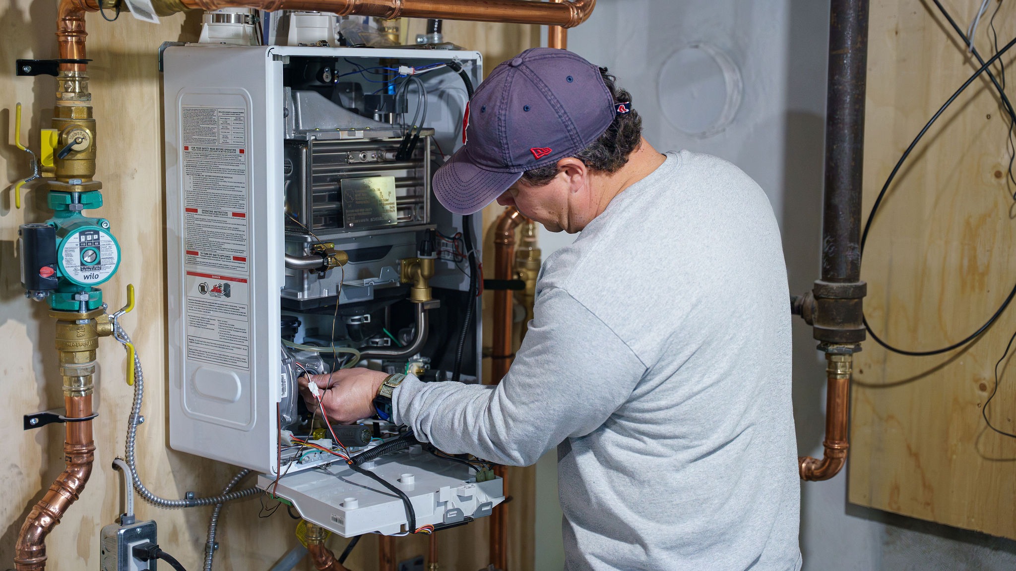 A person wearing a cap works on a wall-mounted boiler system with exposed pipes and wiring, focused on repairs or installation.