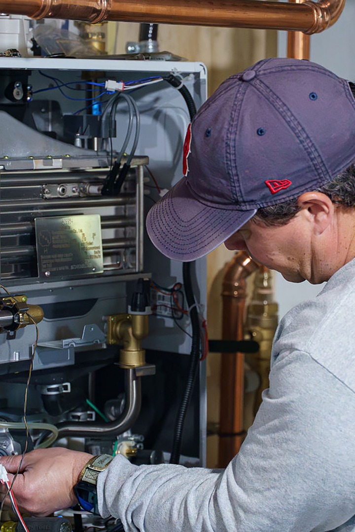 A person wearing a cap is working on intricate machinery with copper pipes, focusing on electrical connections, possibly repairing or installing equipment.