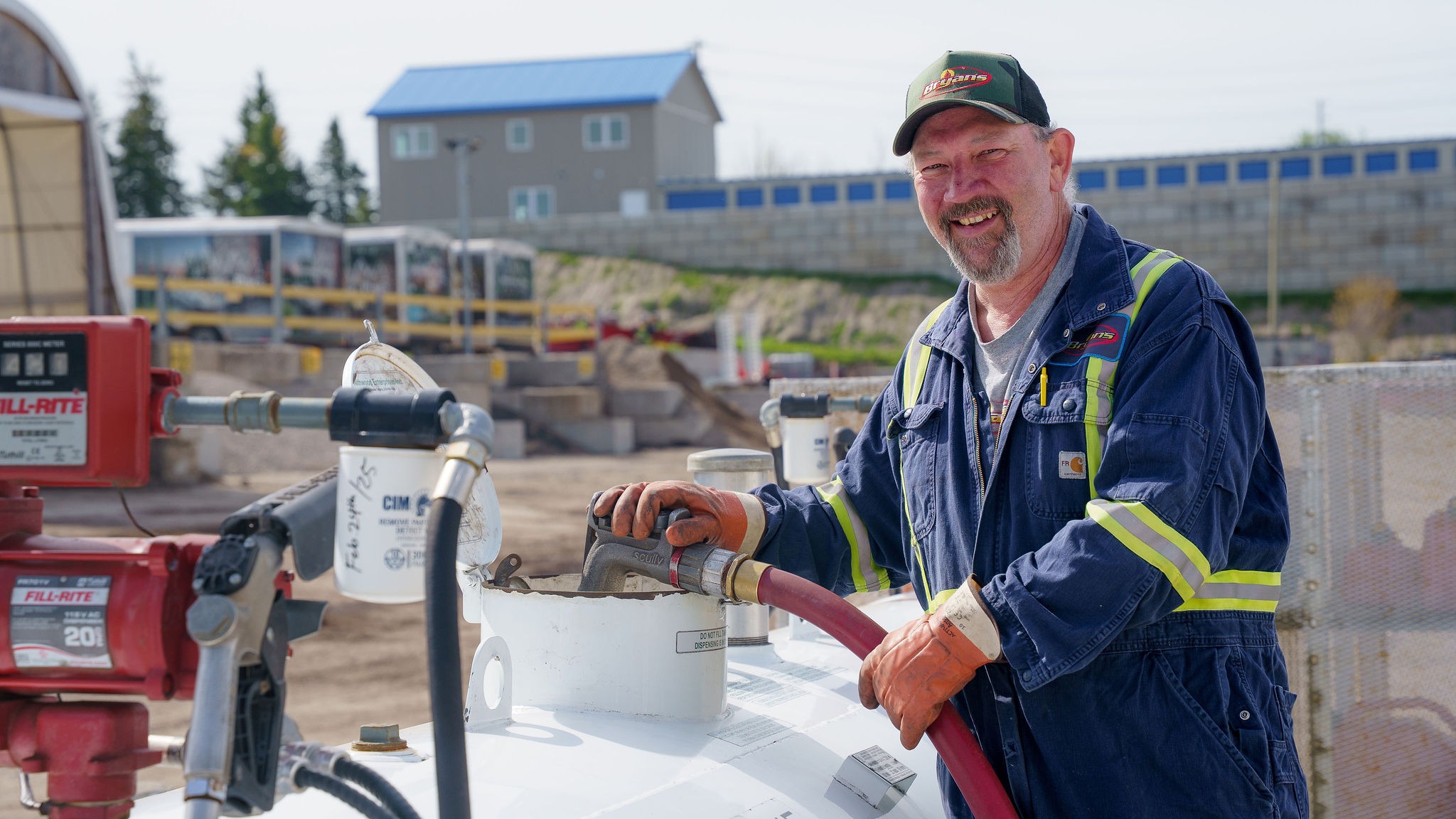 A person in work attire refuels a tank outdoors, smiling. Industrial setting with equipment, bins, and buildings in the background.