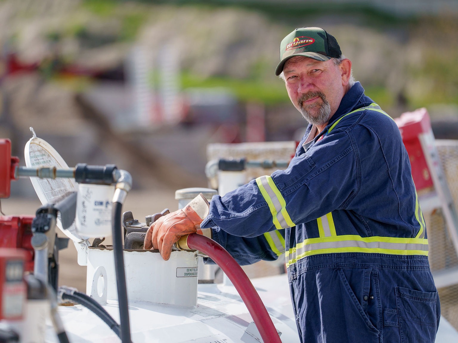 A person wearing protective gear operates machinery outdoors, surrounded by hoses and valves, with an industrial landscape blurred in the background.