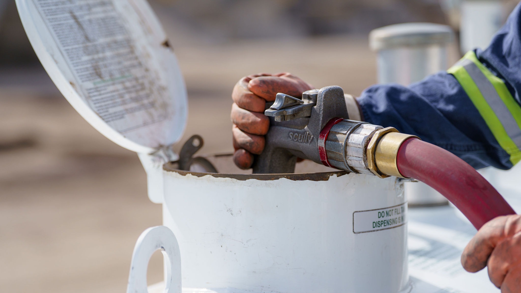 A person operates a fuel nozzle attached to a red hose, filling a white container outdoors, with a safety warning visible nearby.