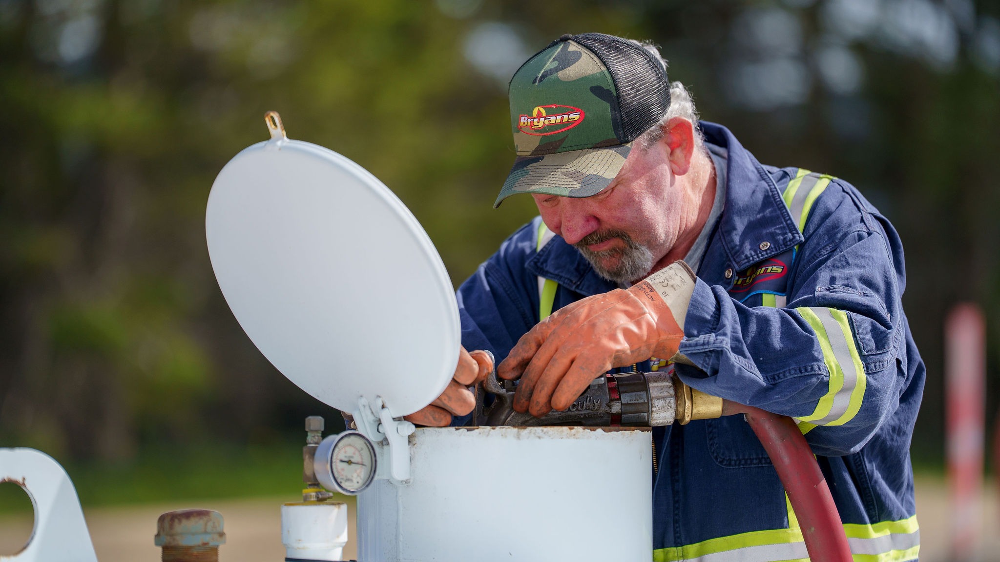 A person in a cap and gloves works on an outdoor tank, focused on adjusting equipment. Background is natural and green.