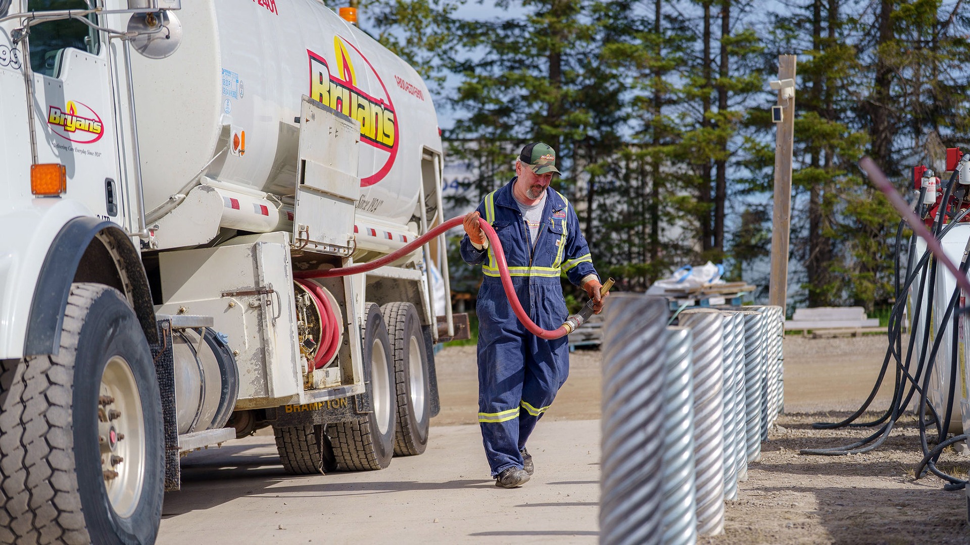 A person in uniform handles a fuel hose from a truck labeled "Bryan's" on a sunny day, with trees in the background.