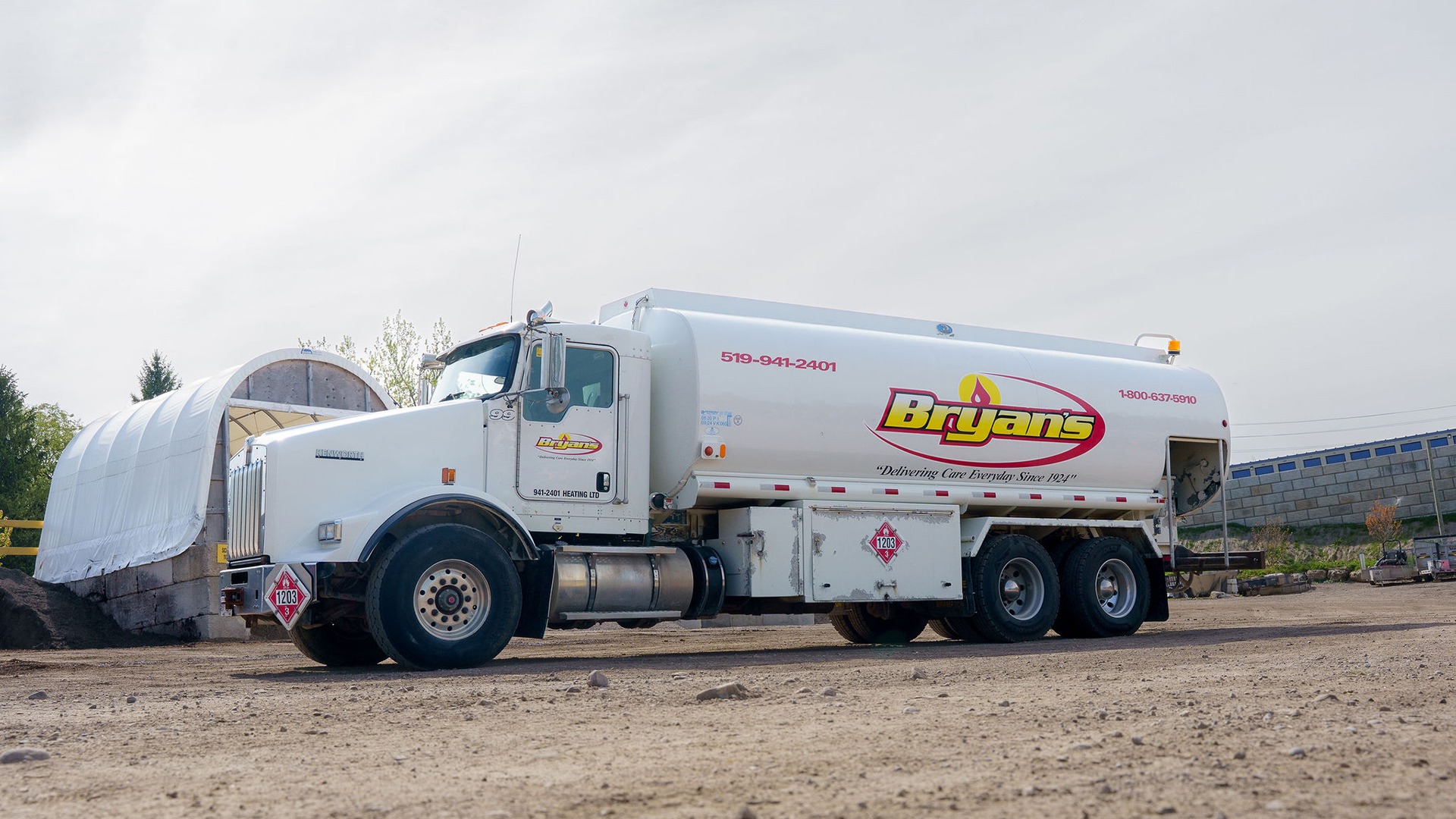 A large white fuel truck with "Bryan's" logo is parked on a gravel surface near an industrial area, with trees in the background.