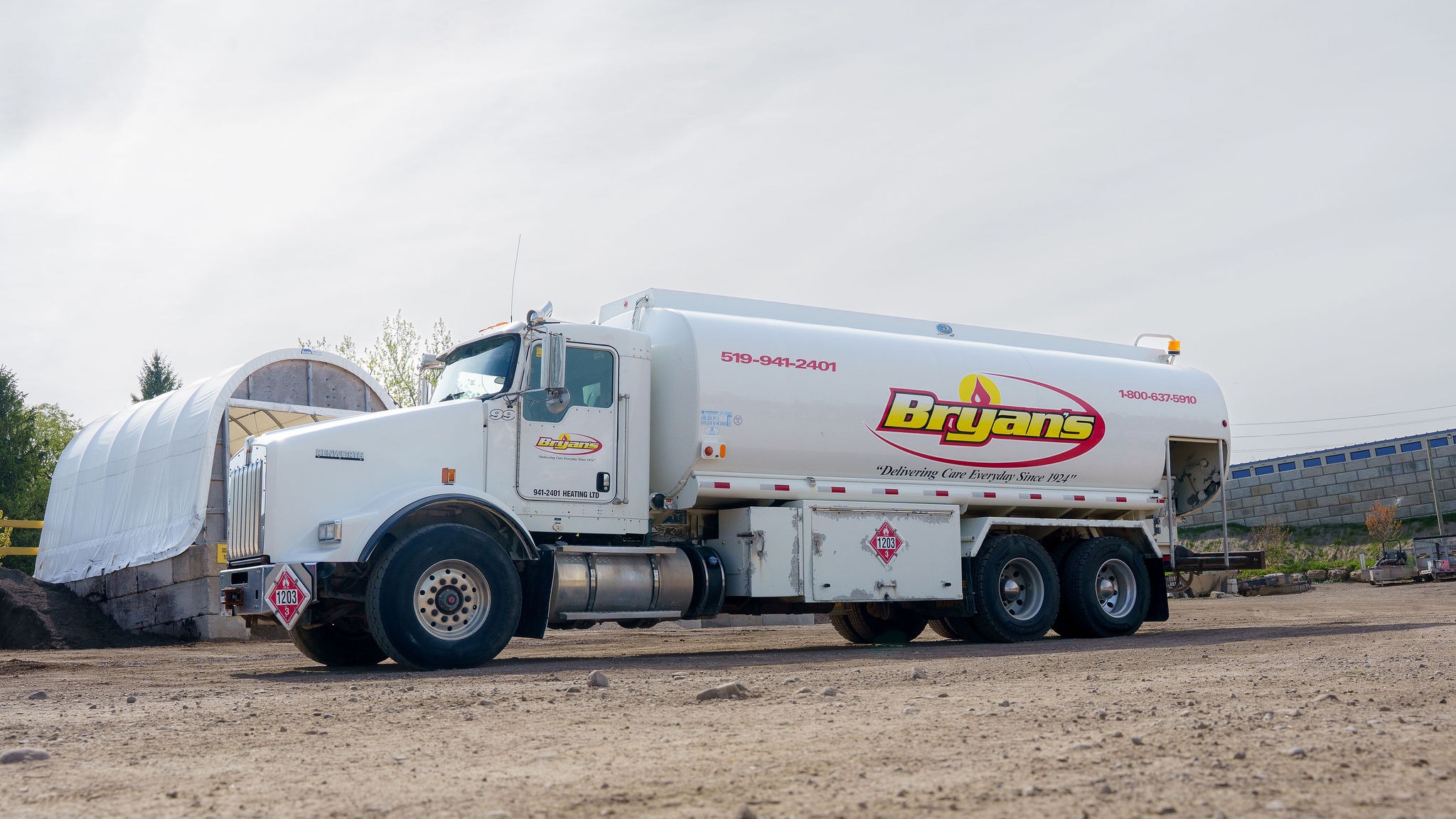 A fuel truck with "Bryan’s" branding is parked on a dirt lot, beside a fabric-covered shelter and a stone building.