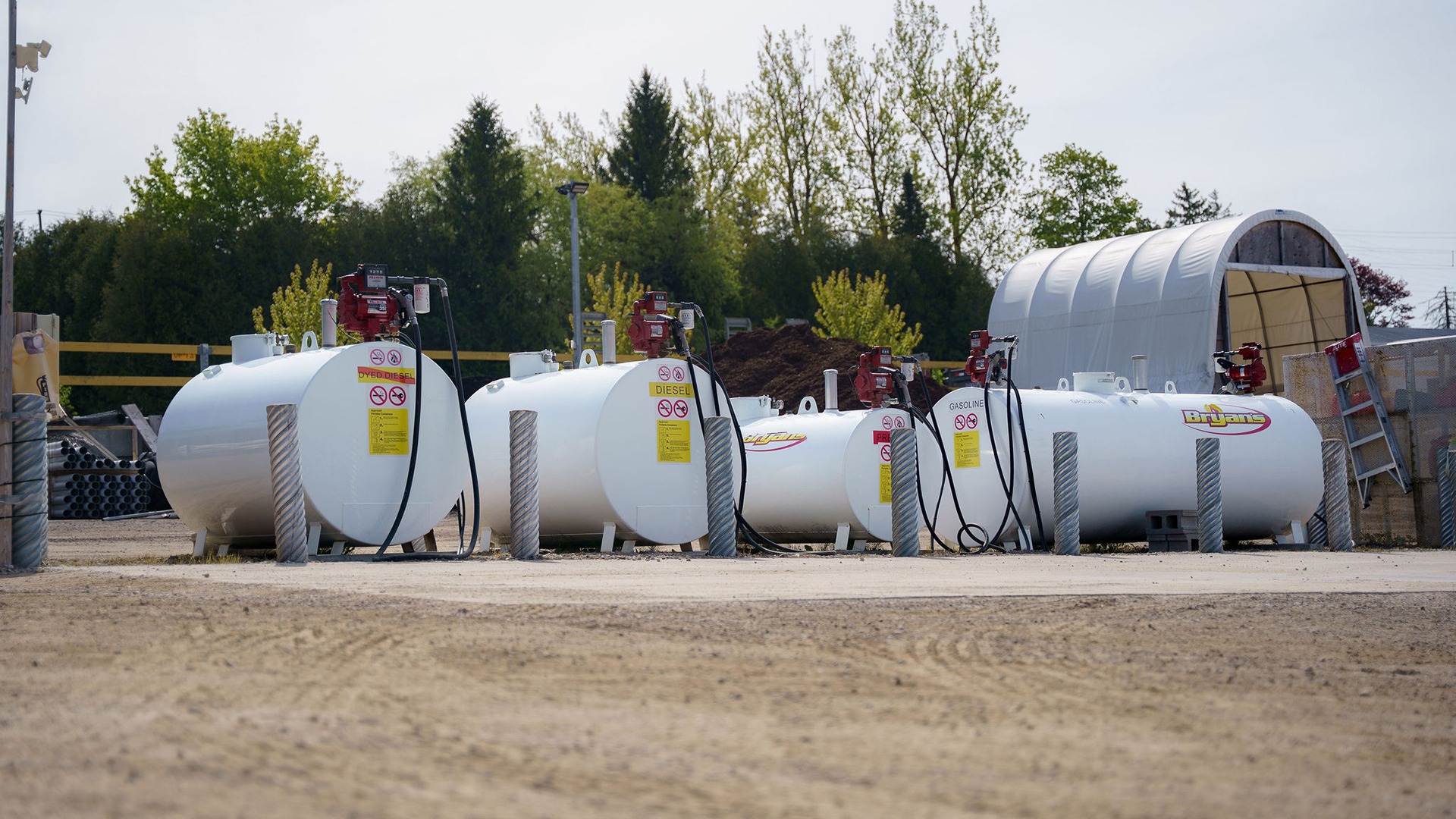 Outdoor scene with multiple white fuel tanks, labeled diesel and gasoline. Surrounded by trees, gravel, and construction materials, with overcast sky above.