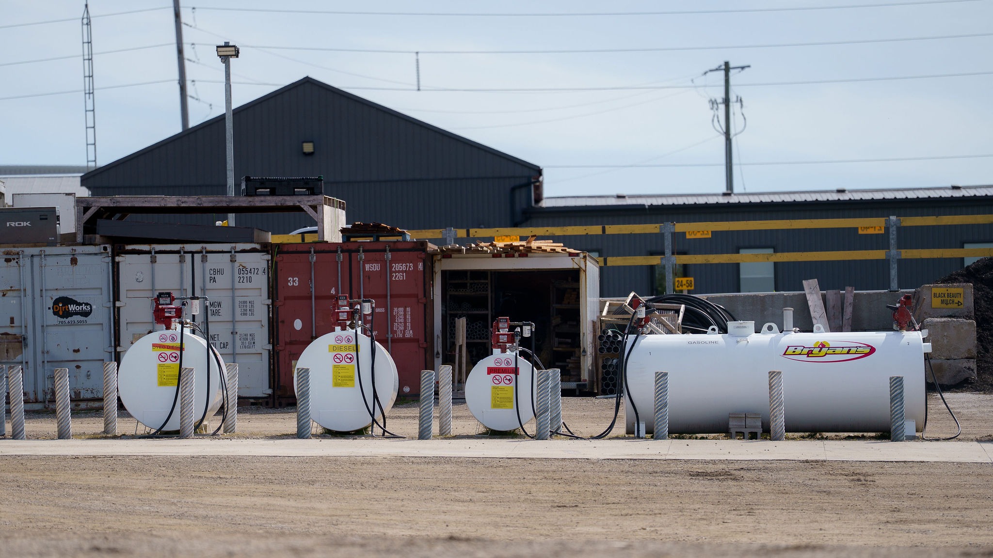 The image shows an industrial site with large fuel tanks, storage containers, and equipment against a backdrop of a warehouse.
