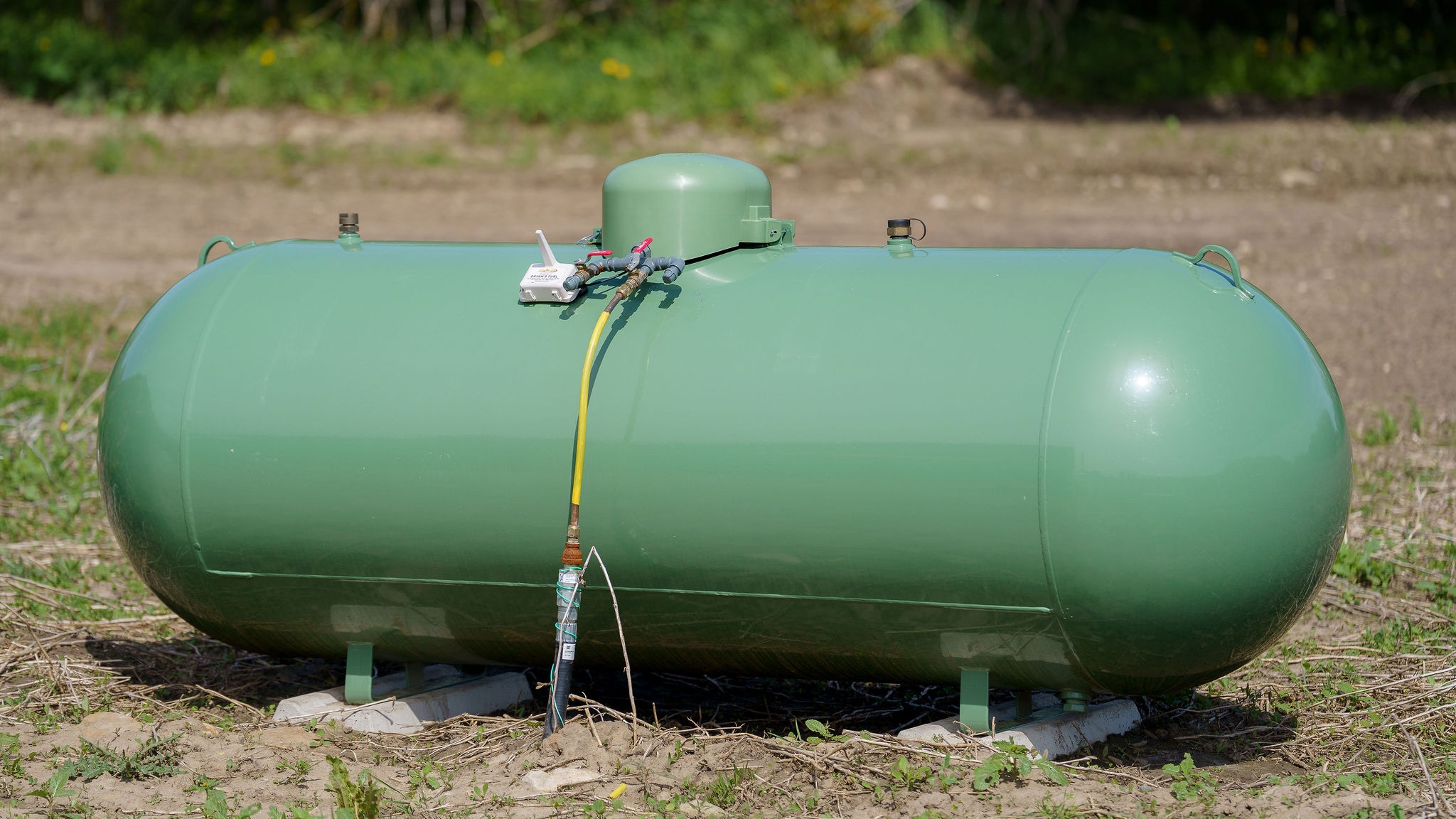 A large green propane tank sits on a grassy field, linked with a yellow hose, under a clear sky, surrounded by vegetation.
