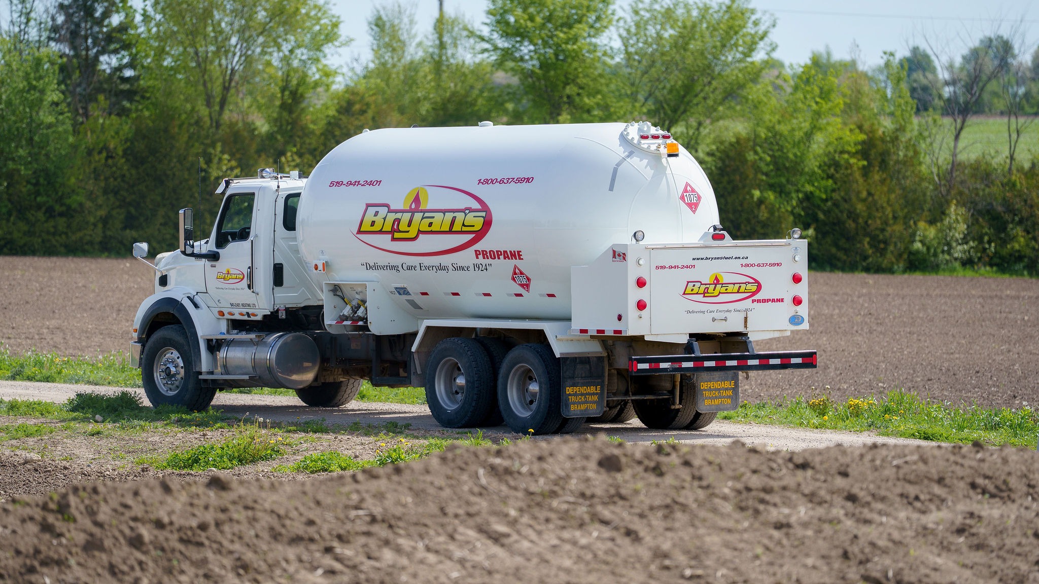 A propane delivery truck drives on a rural road surrounded by fields and trees, under a clear sky. No landmarks or people visible.