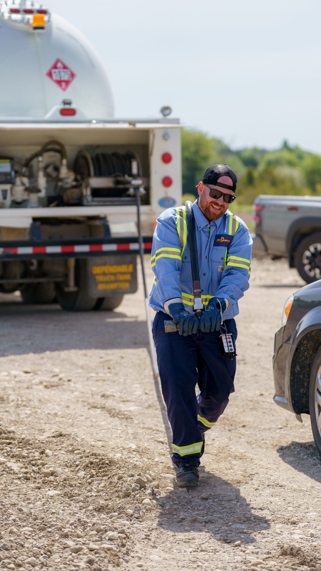 A person in a safety uniform pulls a fuel hose on a dirt road beside a truck, wearing sunglasses and a cap, under clear skies.