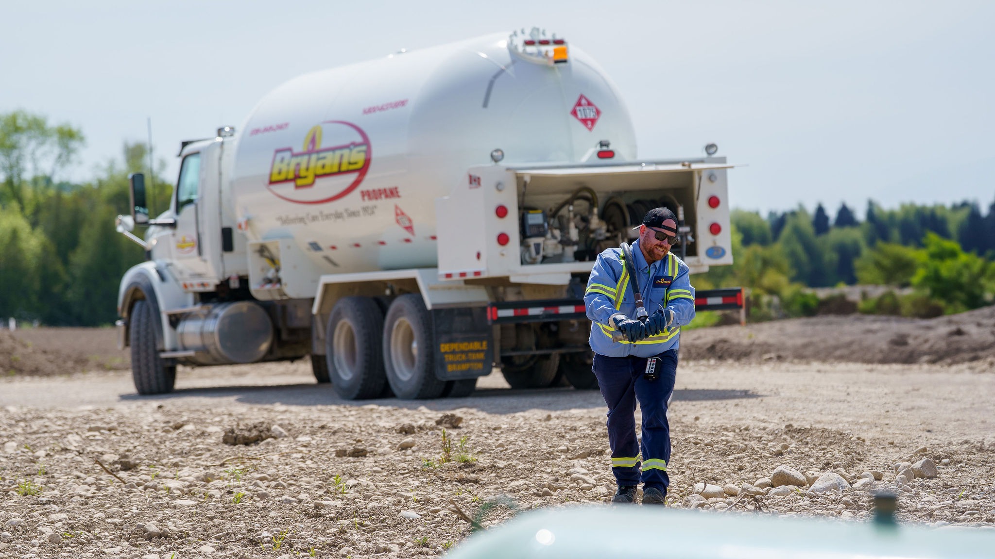 A person works outdoors near a propane truck on a rocky path, surrounded by trees and open land.