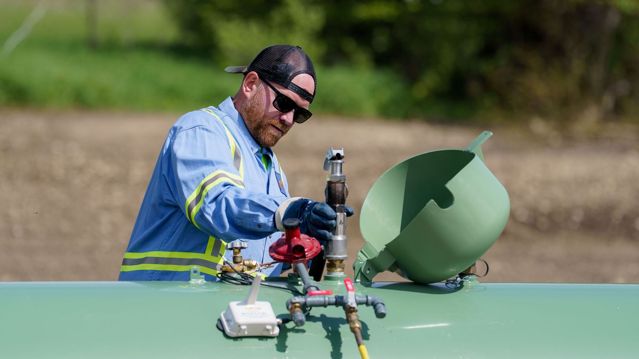 A person in safety gear works on a gas pipeline outdoors, adjusting valves and wearing sunglasses, with greenery in the background.