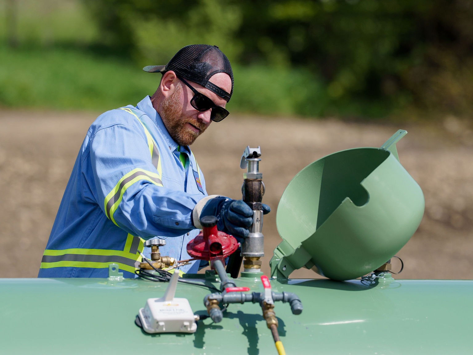 A person wearing safety gear works on green equipment outdoors, possibly related to utilities or construction, with a blurred natural background.