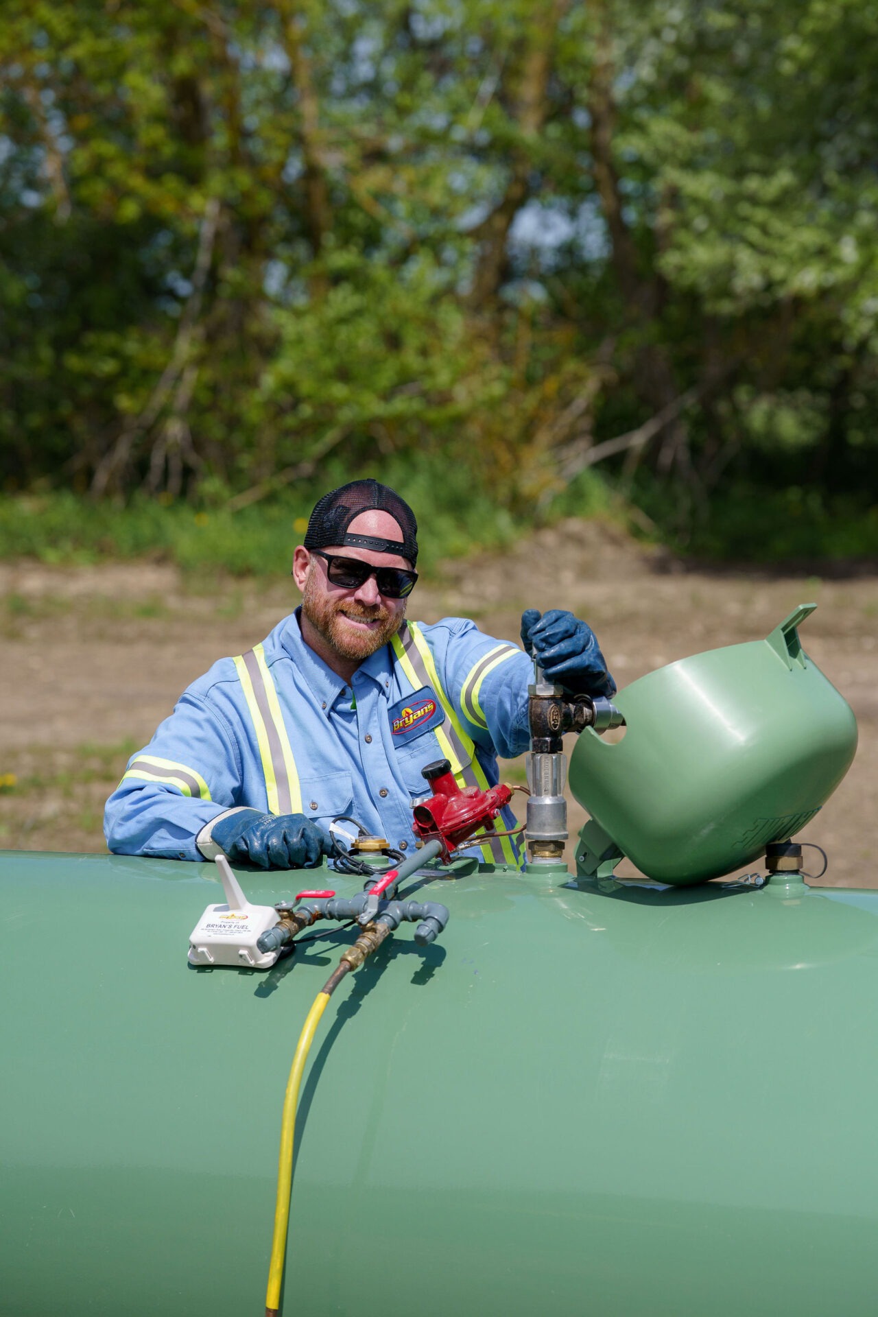 A person wearing safety gear inspects equipment on a large green tank outdoors, surrounded by trees and a natural setting.