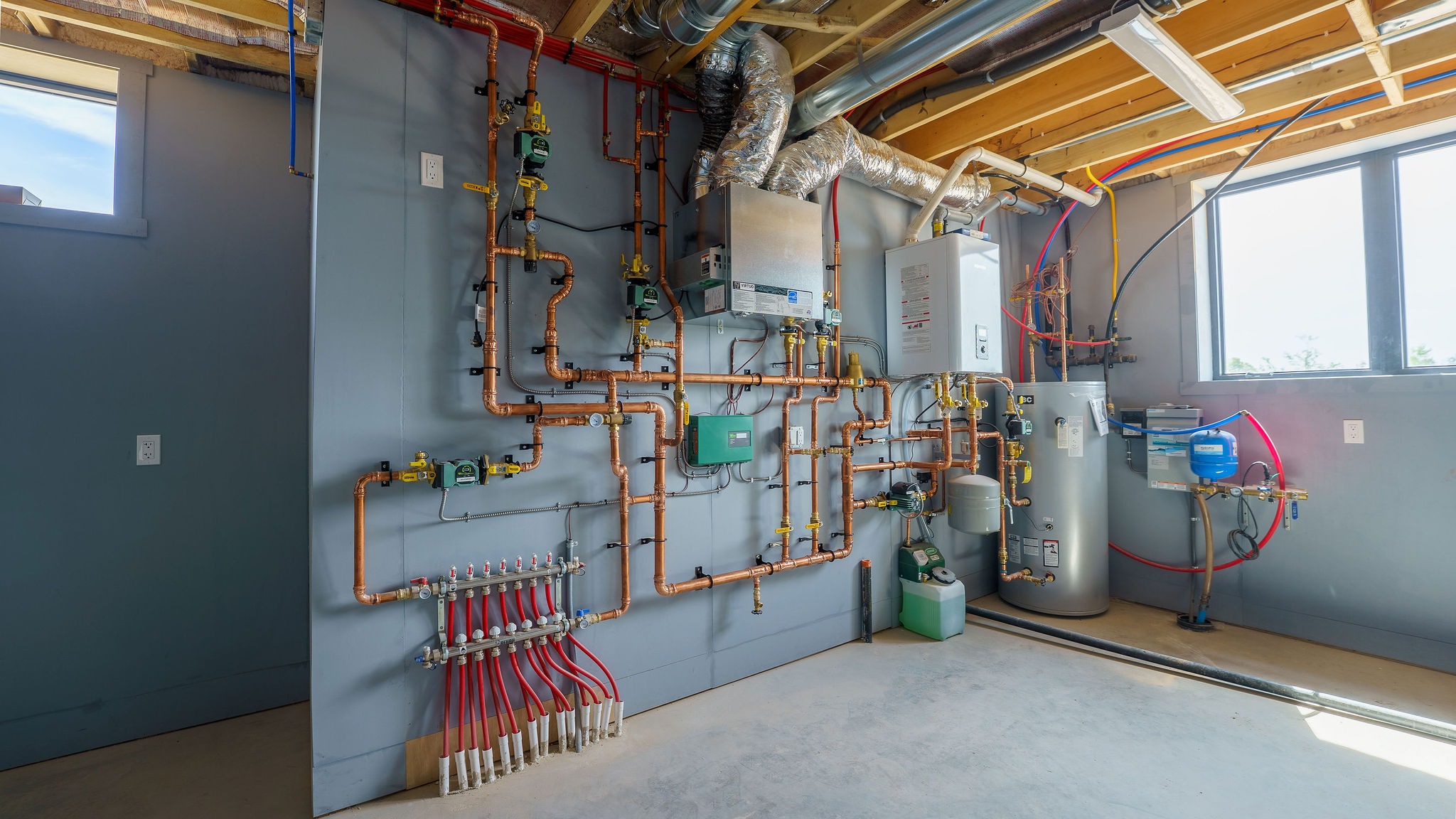 Complex heating and plumbing system with copper pipes, tanks, and valves in a bright utility room, well-organized against a gray wall.