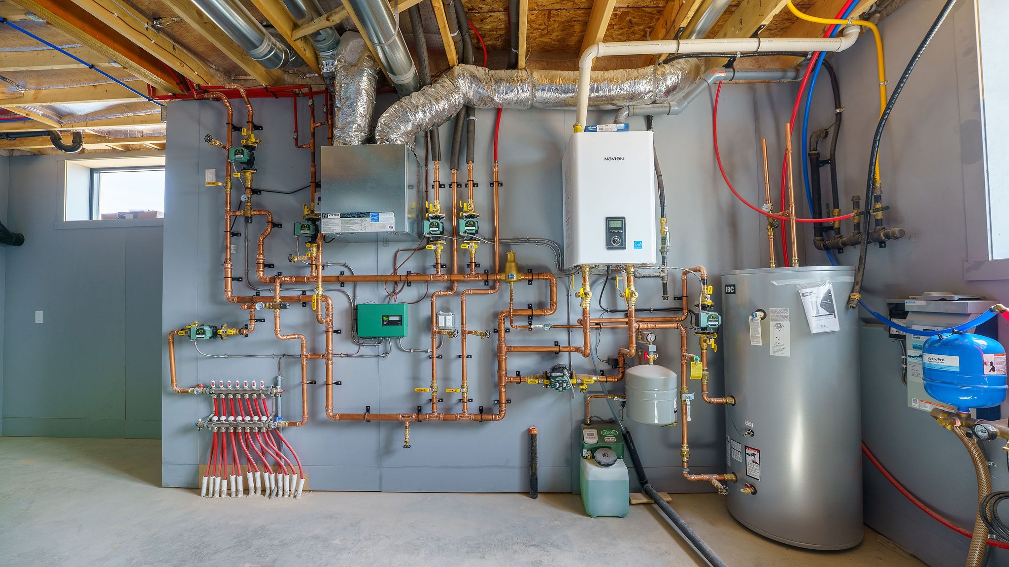 Mechanical room with pipes, tanks, and HVAC equipment neatly organized on a gray wall. Exposed ceiling shows various ducts and wires. Bright lighting.