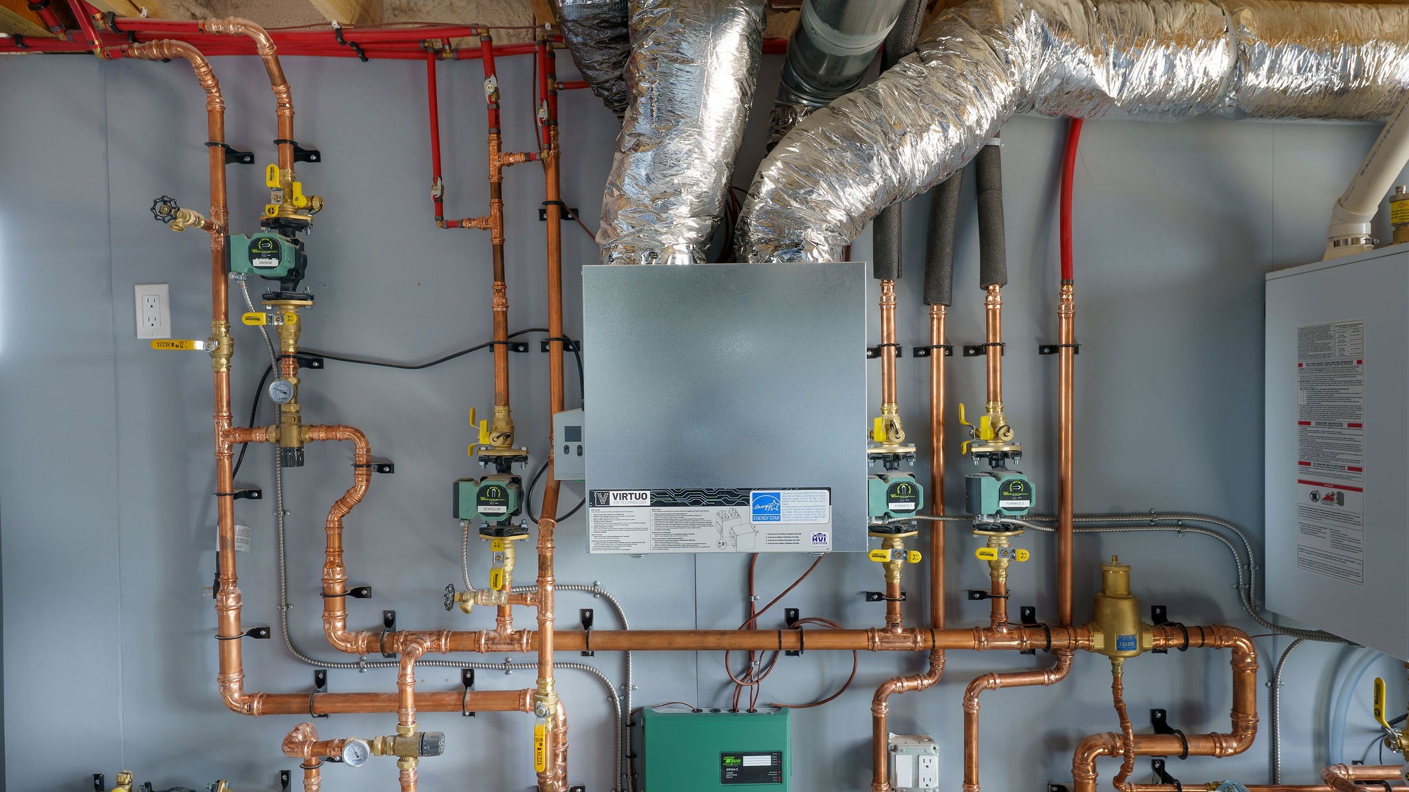 Complex plumbing system with copper pipes, valves, and insulated ducts connected to a heating unit on a wall. No people or recognizable landmarks.