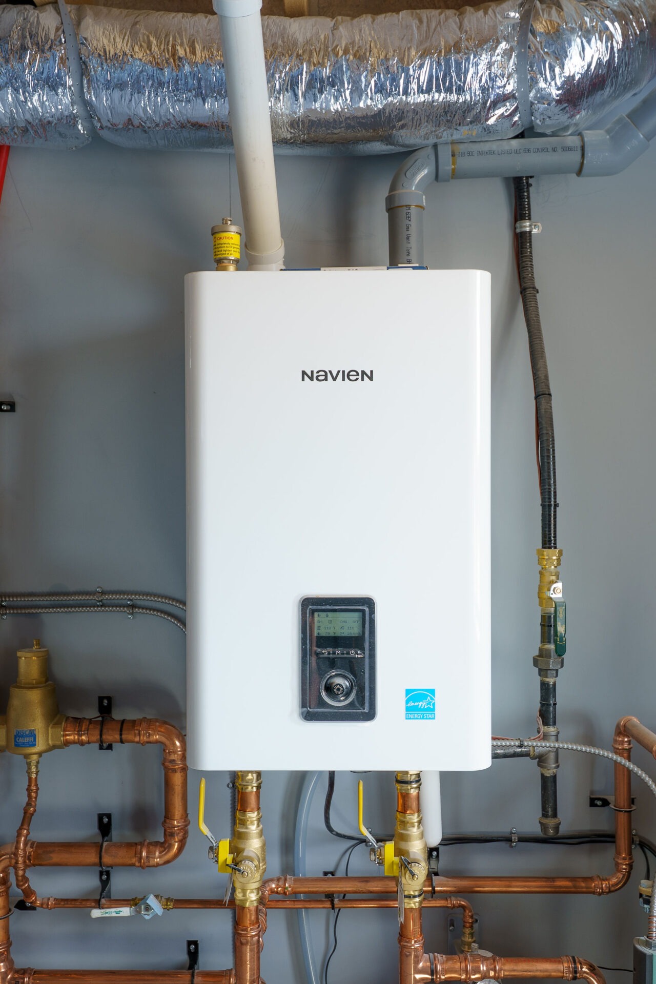 A white Navien tankless water heater with copper piping attached, installed on a wall, with ductwork and other plumbing visible.