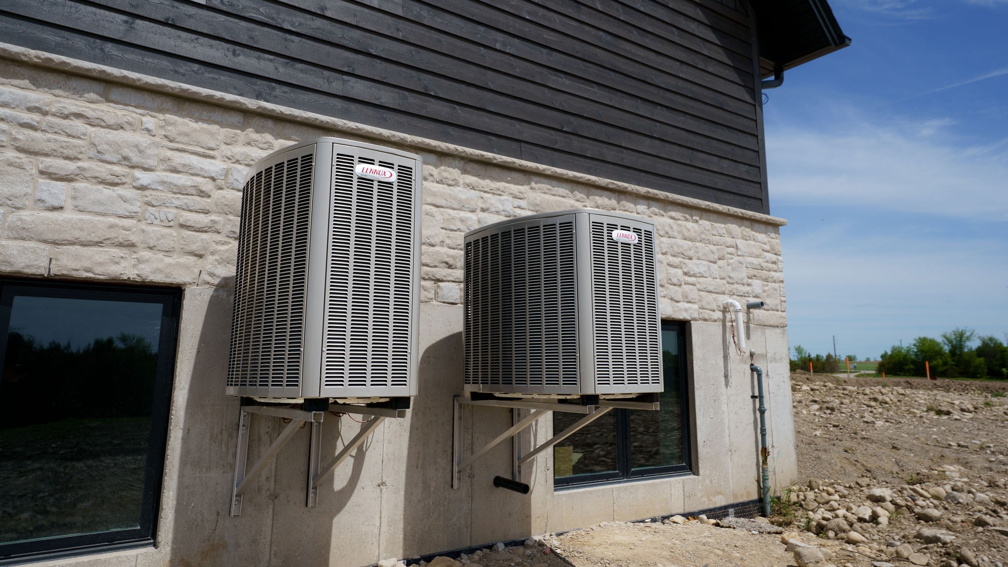 Two outdoor air conditioning units mounted on a stone wall of a building, with a dirt ground and blue sky in background.