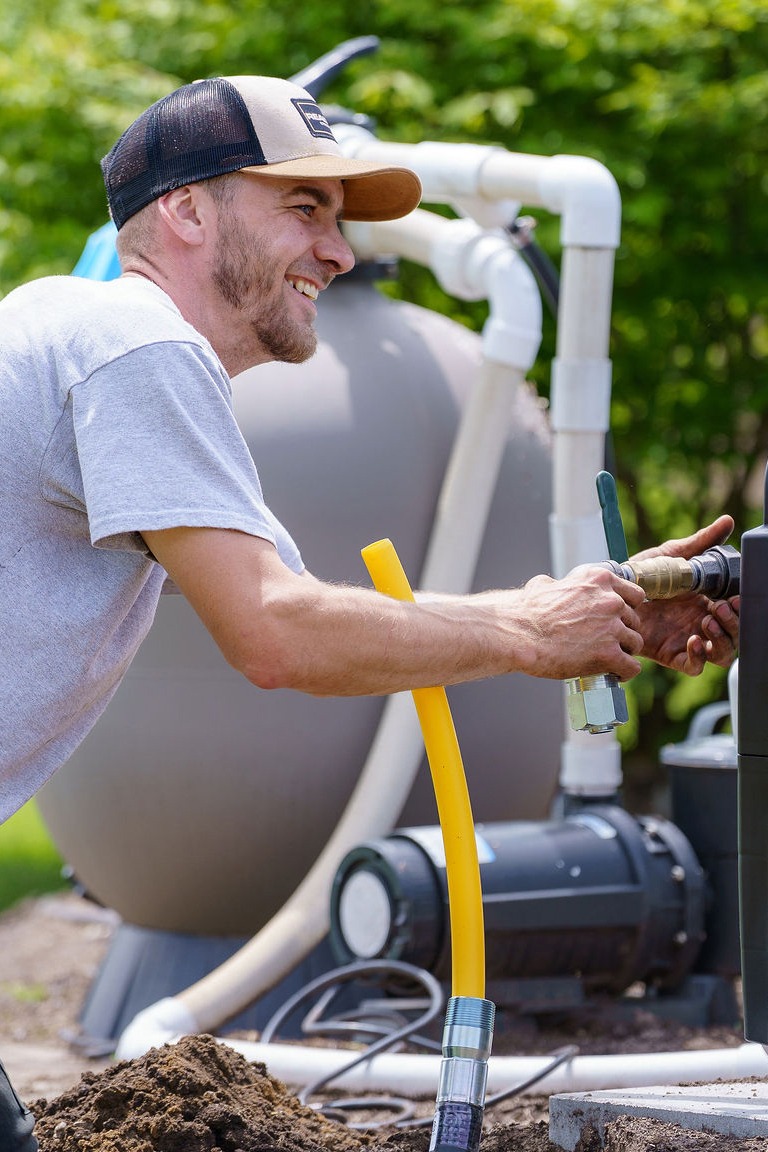A person wearing a cap smiles while working with pipes outdoors. Equipment and lush greenery are visible in the background.