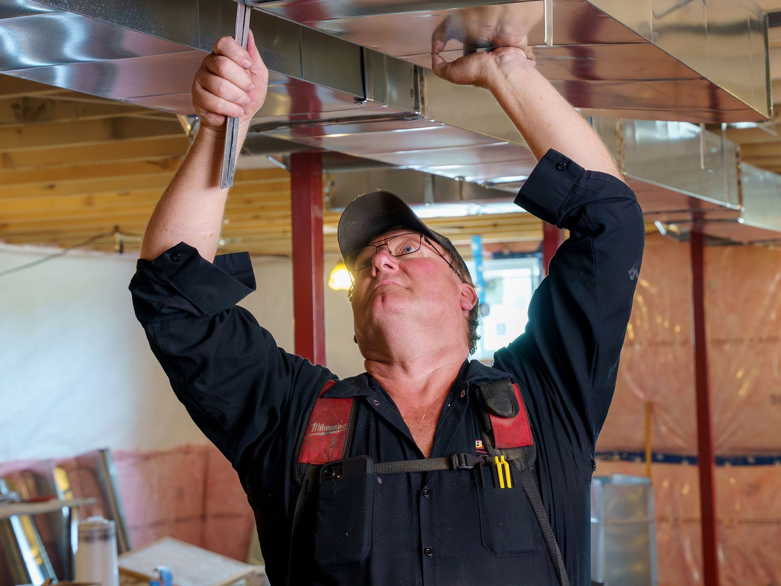 A person in a cap is installing metal ductwork on a ceiling in a construction site, using tools to secure components.
