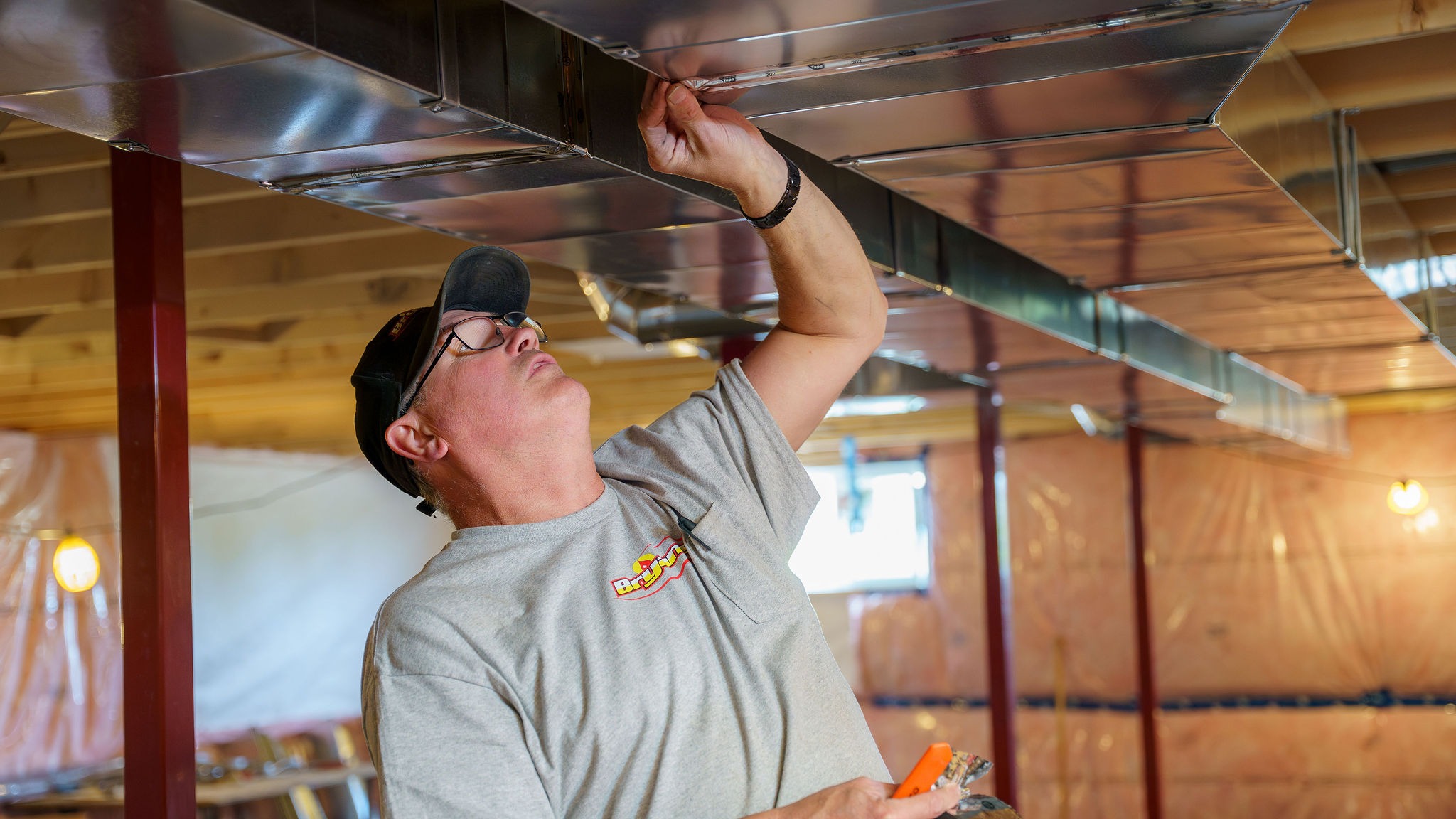 A person is working on duct installation in a partially constructed interior space, wearing a gray shirt and cap, holding a tool.