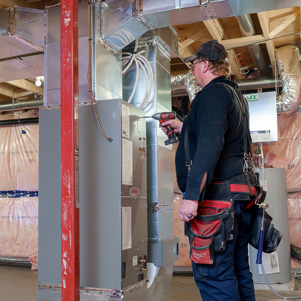 A person in a basement is using a drill on a heating unit, surrounded by ductwork and insulation material.