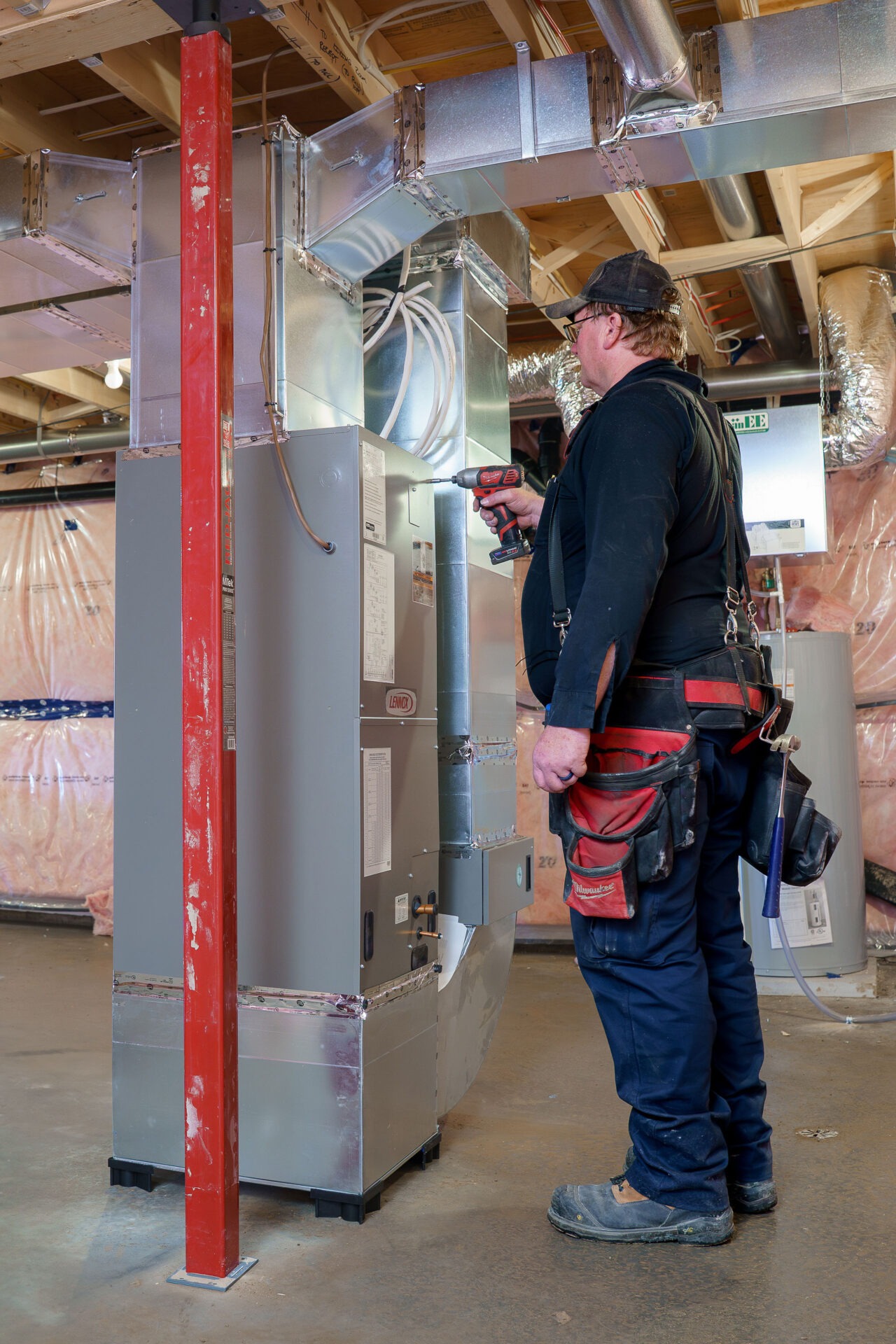 A person in a basement installs HVAC ductwork, holding a drill, wearing a toolbelt, surrounded by insulation and exposed beams.