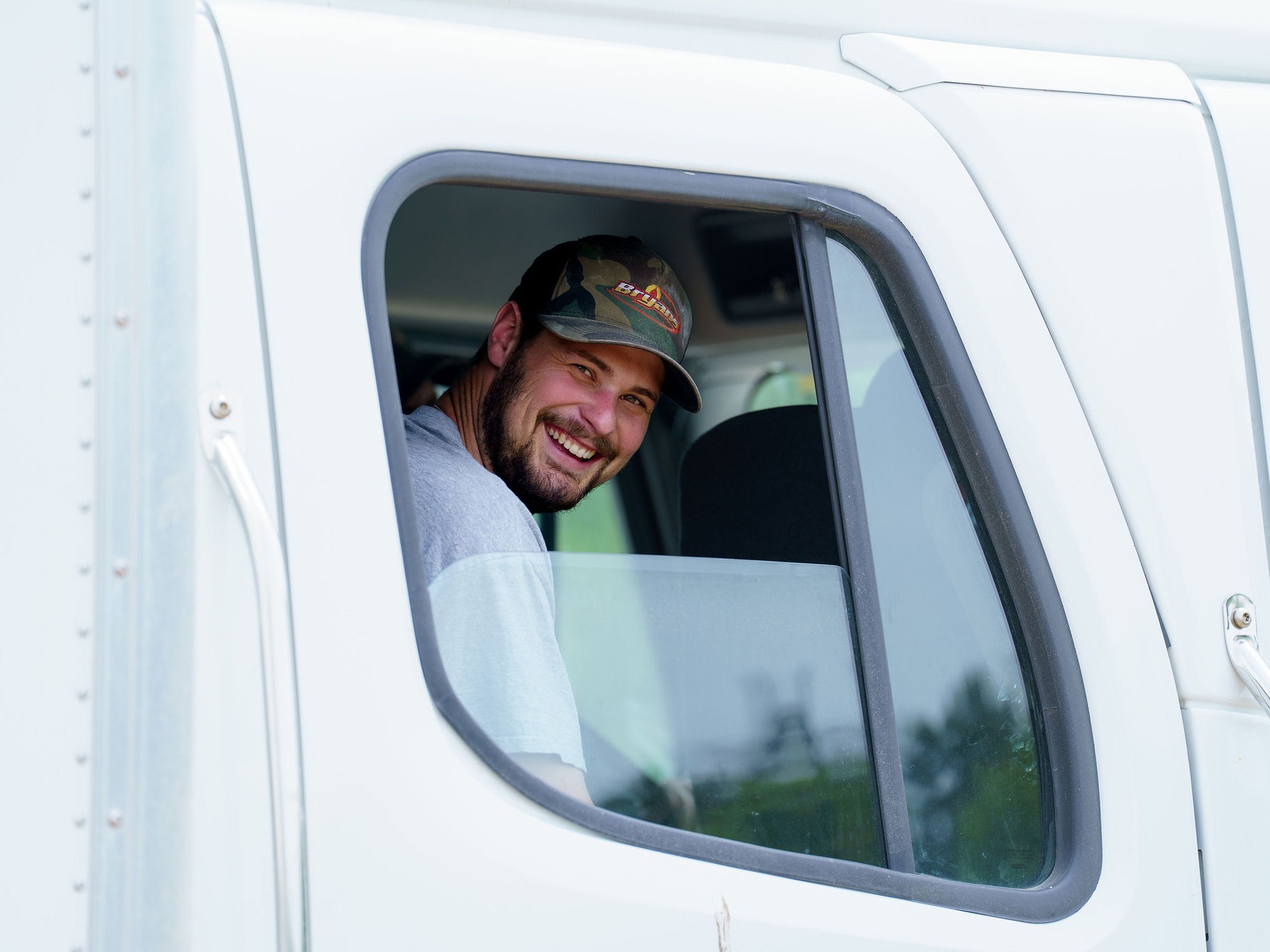 A smiling person wearing a cap looks out the window of a white truck cab, with greenery visible outside.