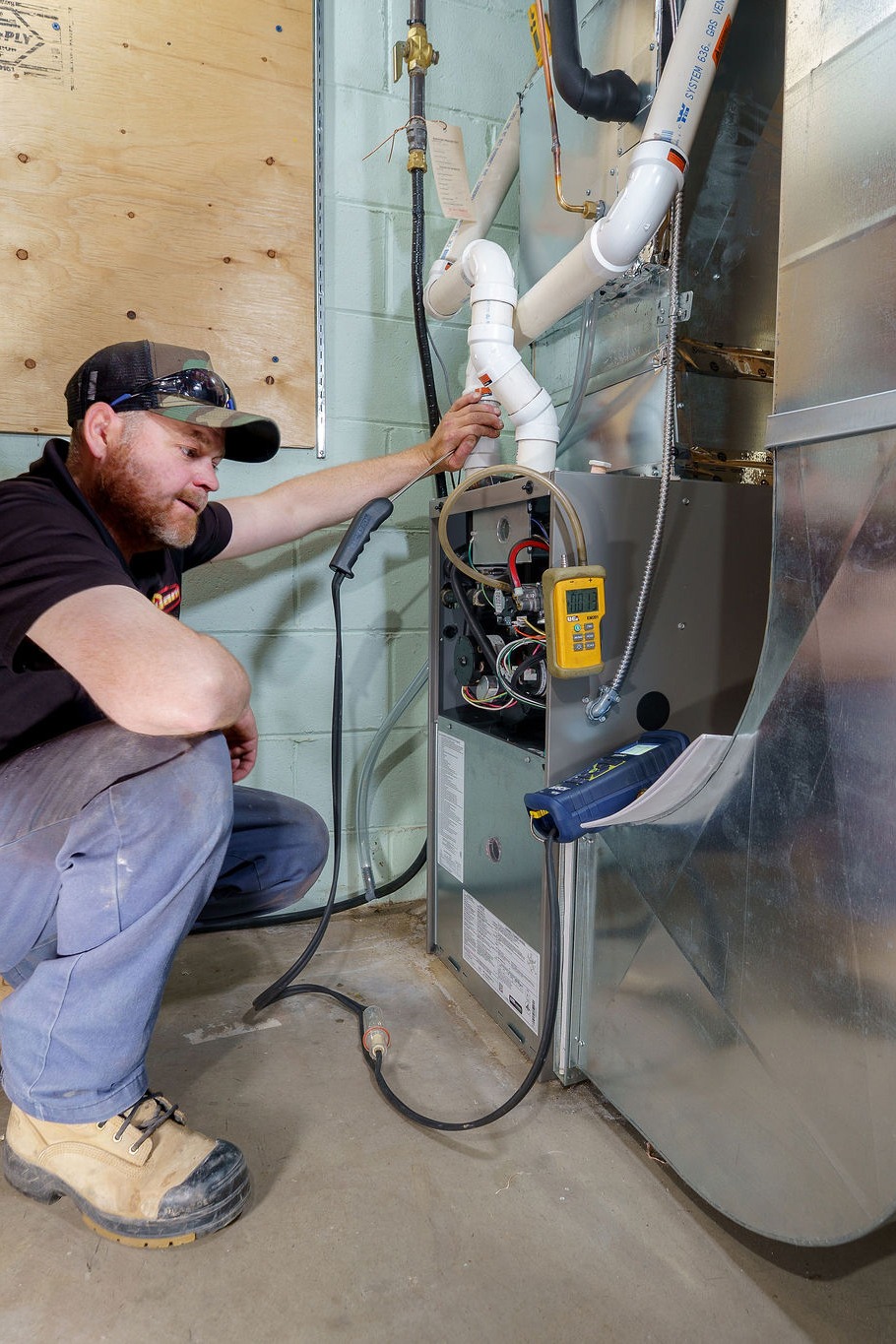 A person is examining and adjusting a furnace system in a utility room, using tools and gauges for maintenance.