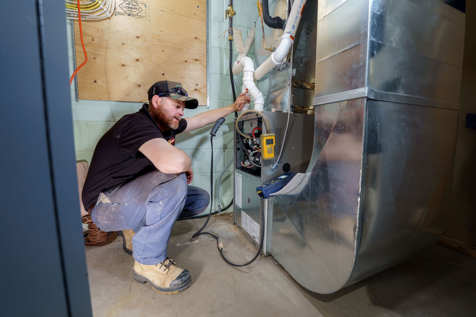 A person wearing work boots examines a furnace system, adjusting components and using diagnostic tools in a utility room.