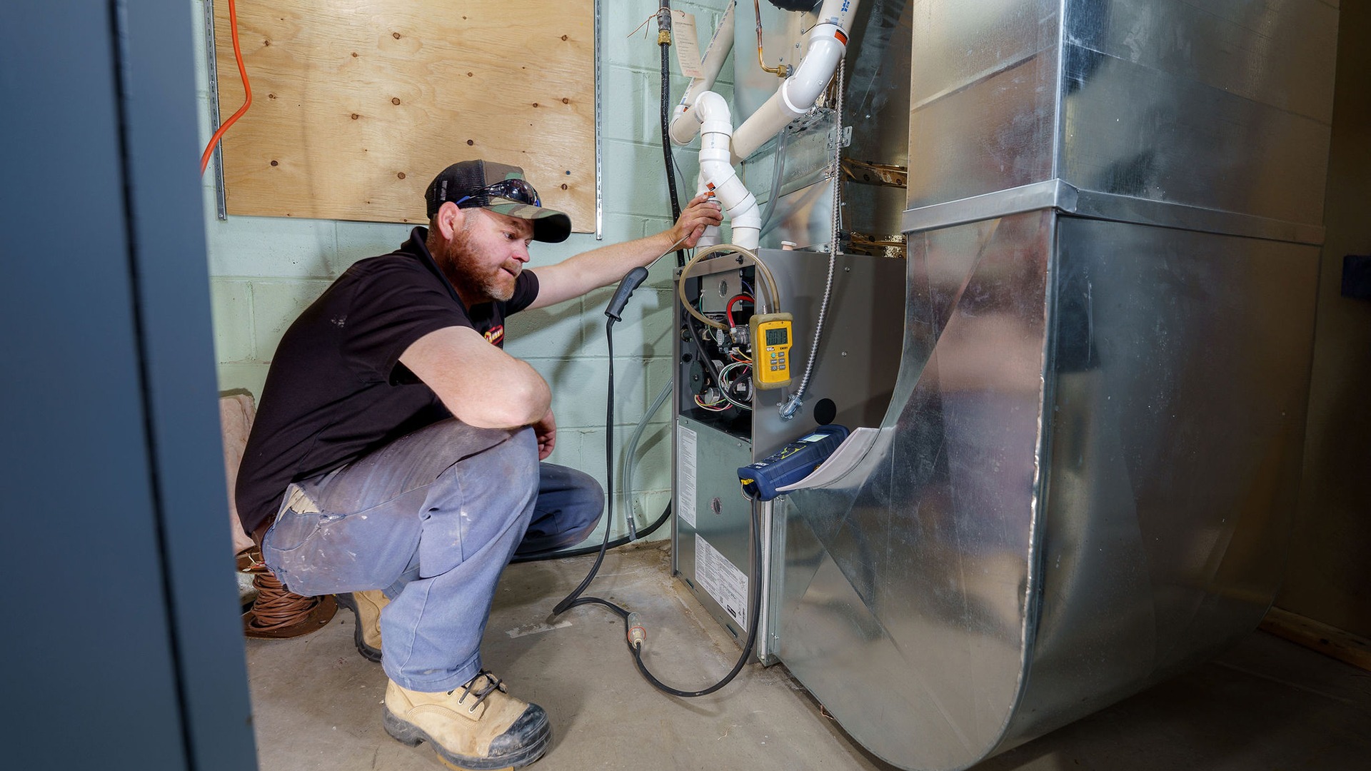 A person kneels, inspecting a furnace in a utility room, surrounded by tools and ducts. No recognizable landmarks or historical buildings.