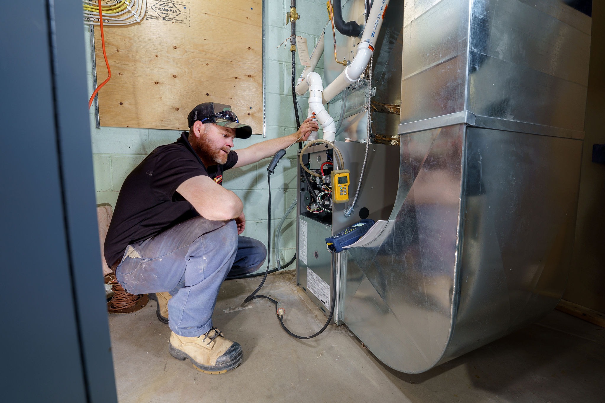 A person wearing work boots examines a furnace system, adjusting components and using diagnostic tools in a utility room.