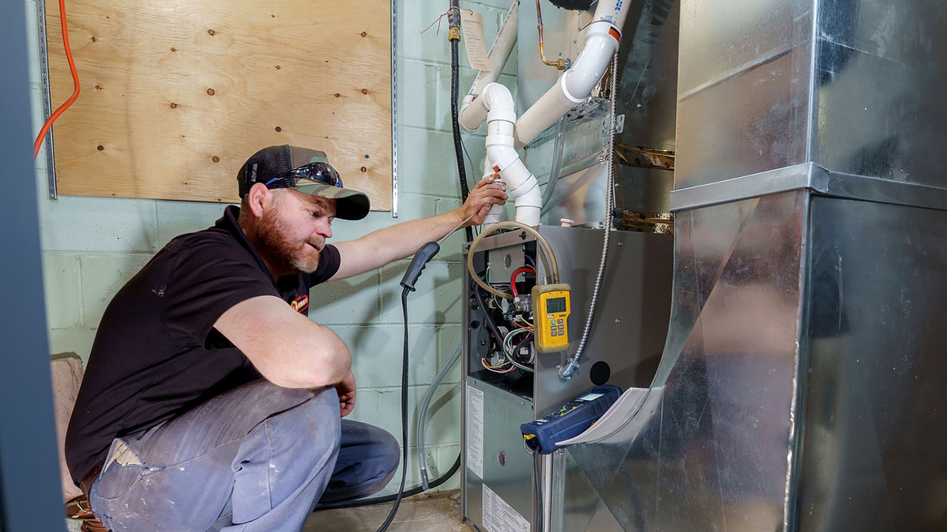 A person in a cap inspects a HVAC system inside a small room with painted cinder block walls and metallic ducts.
