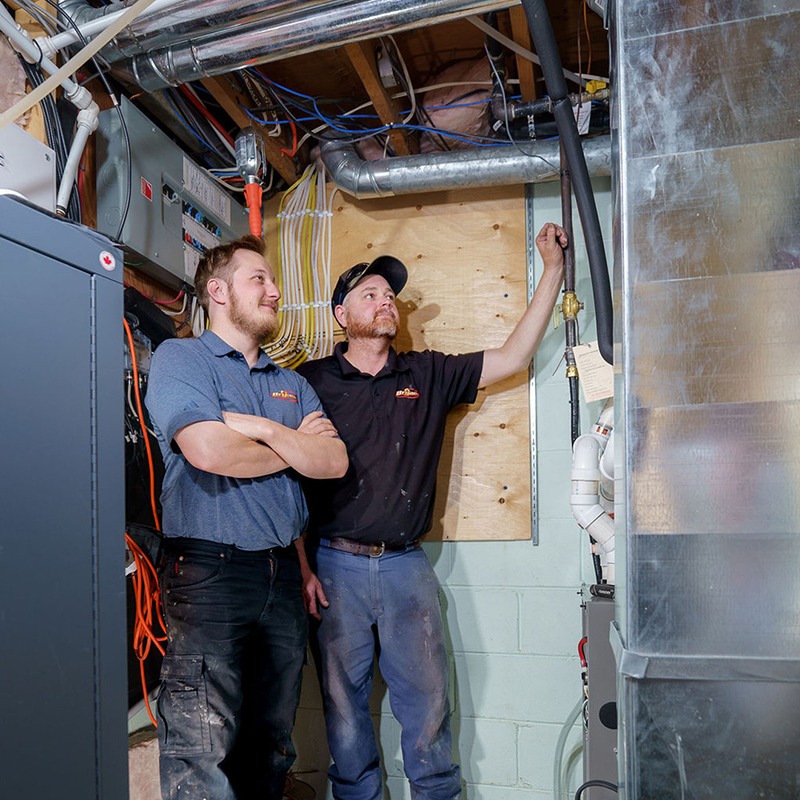 Two people in a utility room, inspecting electrical and plumbing systems, with pipes and wires visible on the wall and ceiling.