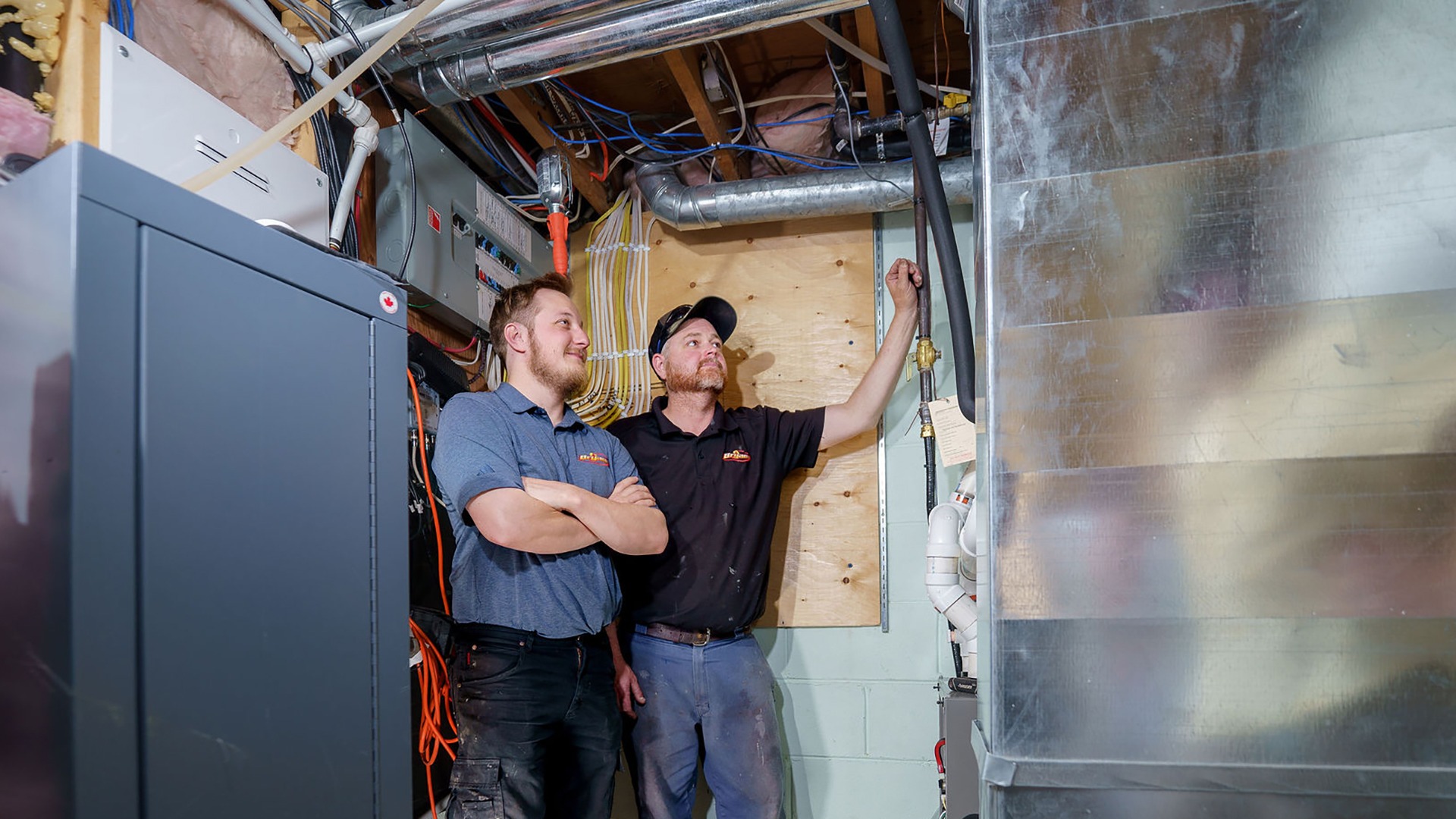 Two people stand in a utility room, inspecting pipes and equipment, surrounded by wires and ducts, focused on a task.