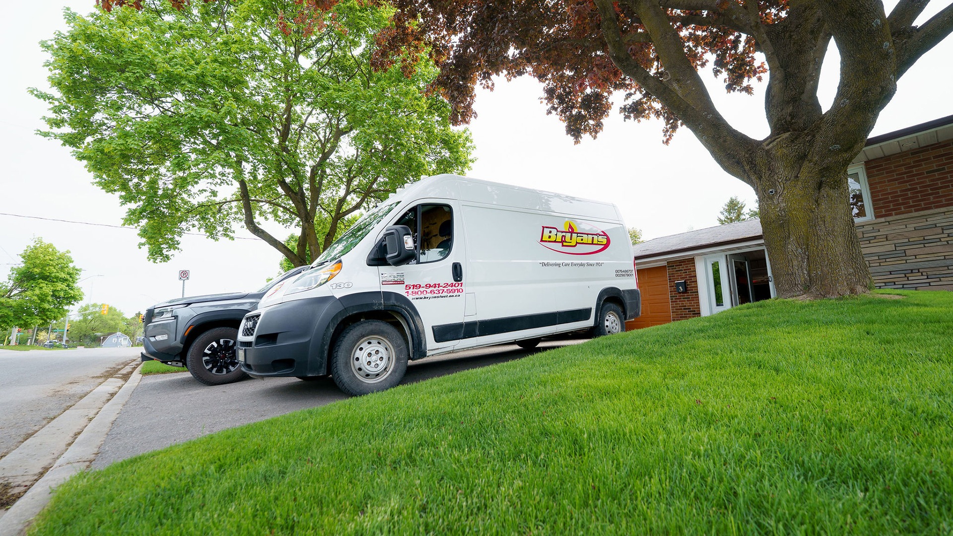 A delivery van parked on a residential street near a house, surrounded by green grass and large trees.