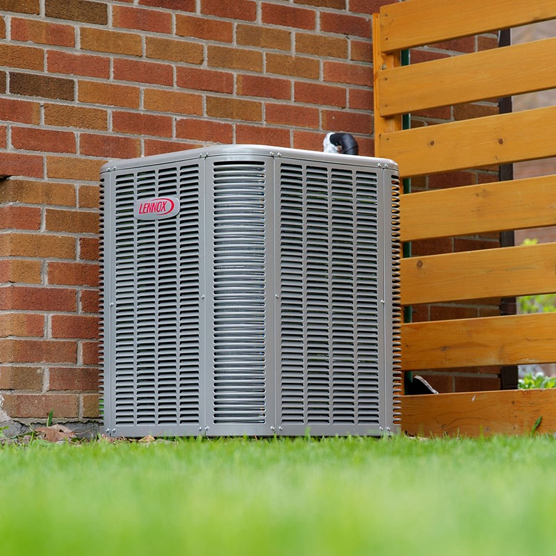An air conditioning unit is situated outside against a brick wall and wooden fence, surrounded by a grassy area.