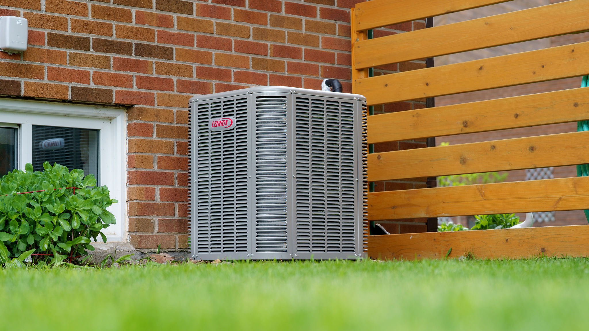 Air conditioning unit outside brick house, near wooden fence and window. Green shrubbery and grass in foreground, clear blue sky visible.