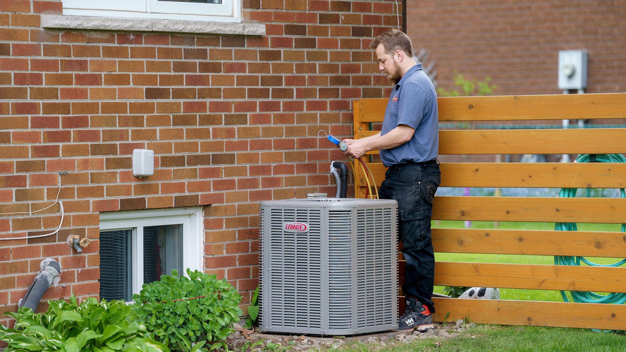 A person checks an air conditioning unit outside a brick house near a wooden fence, surrounded by green plants and a garden hose.