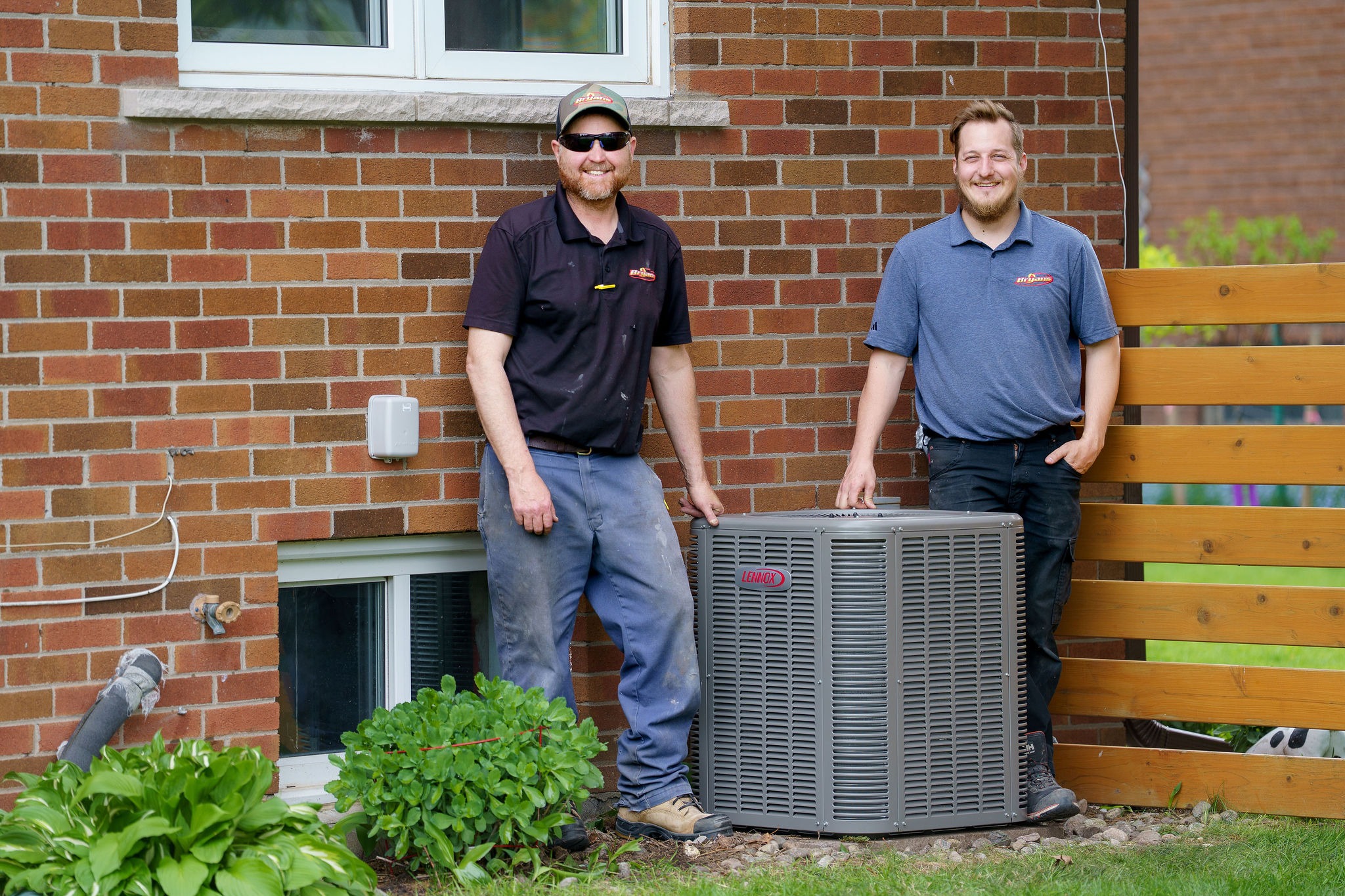Two people smiling beside a Lennox air conditioning unit, with a brick house background. Small window and wooden fence visible.