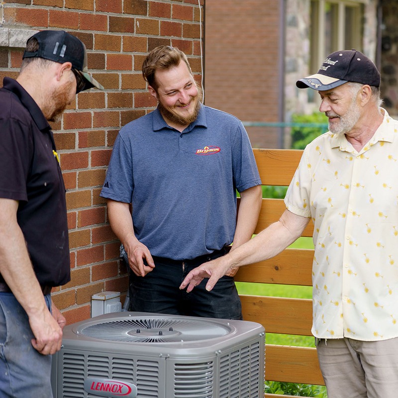 Three people discuss a Lennox air conditioning unit near a brick building. They're wearing casual clothes and seem engaged in conversation.