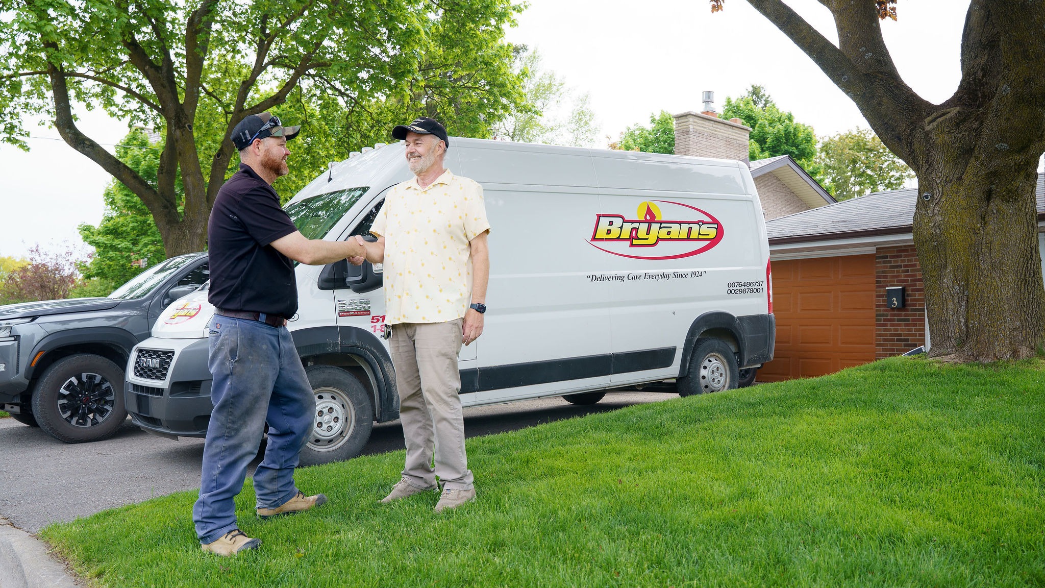 Two people shake hands beside a Bryans' van in a residential neighborhood with trees and brick houses, on a sunny day.