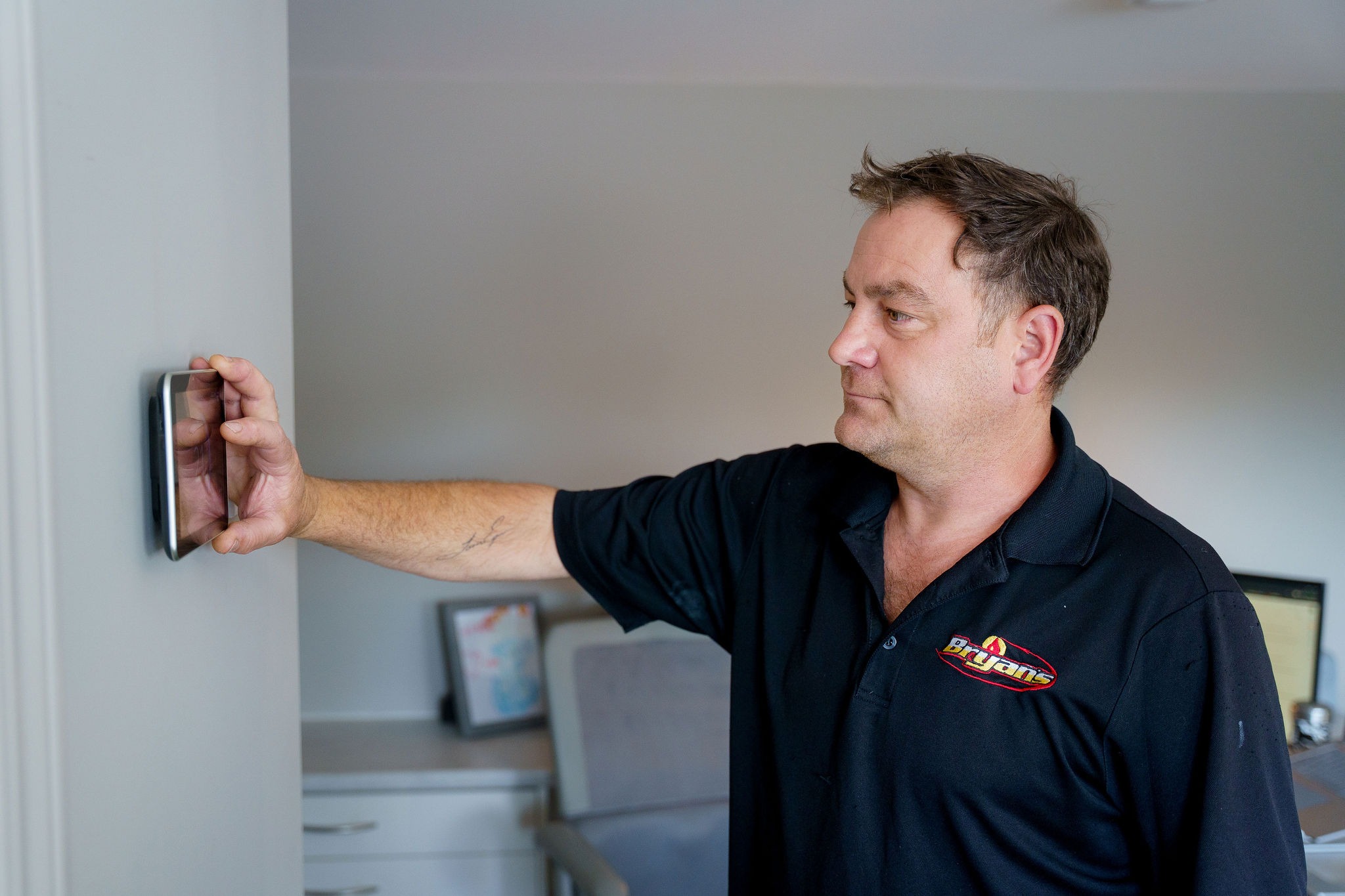 A person in a room adjusts a wall-mounted digital thermostat, wearing a dark shirt. Nearby, a desk with a framed picture.